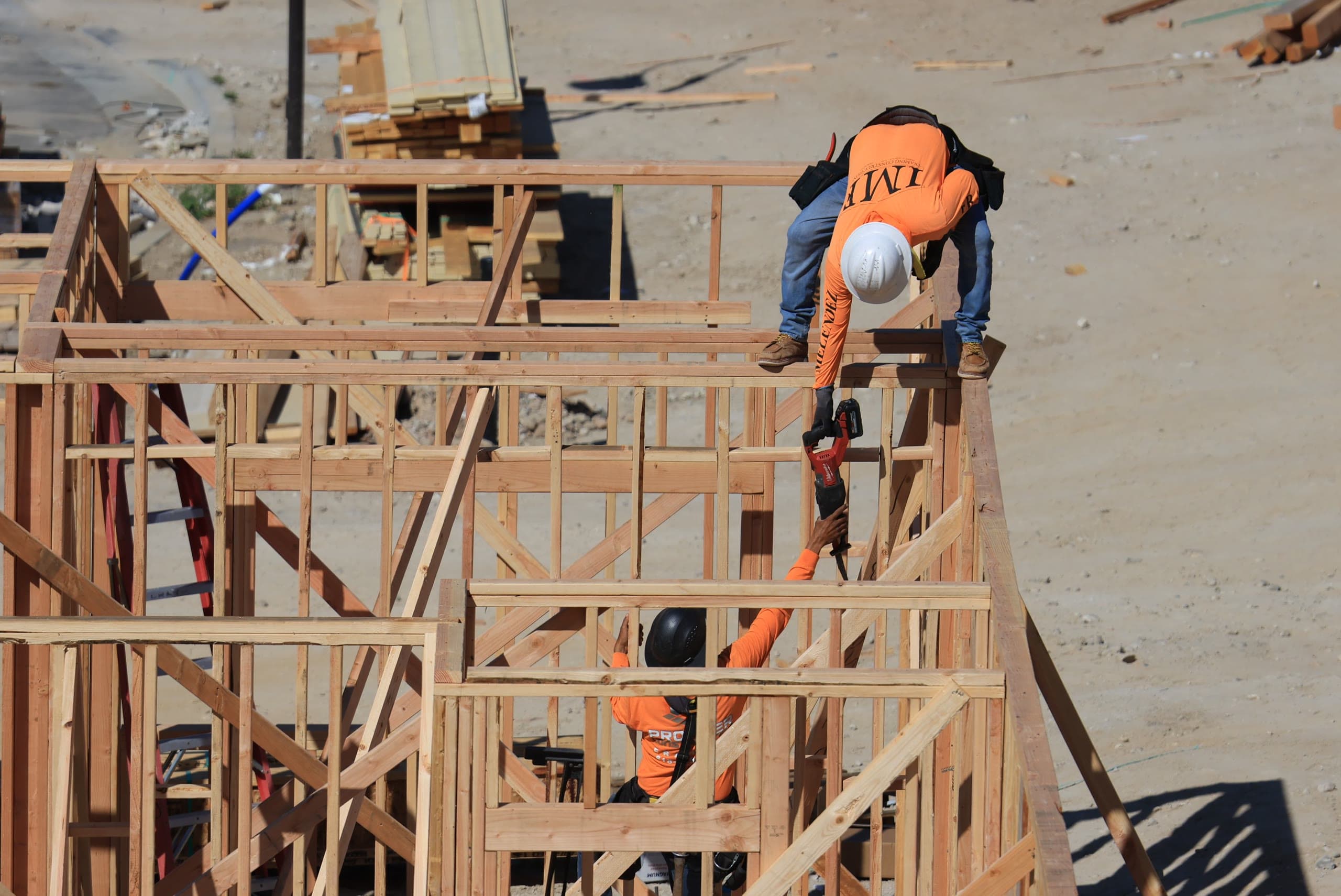 Overhead view of two framing crew members in orange hi-vis shirts and hard hats working on a wood-framed wall, passing a reciprocating saw between the top plate and the floor below