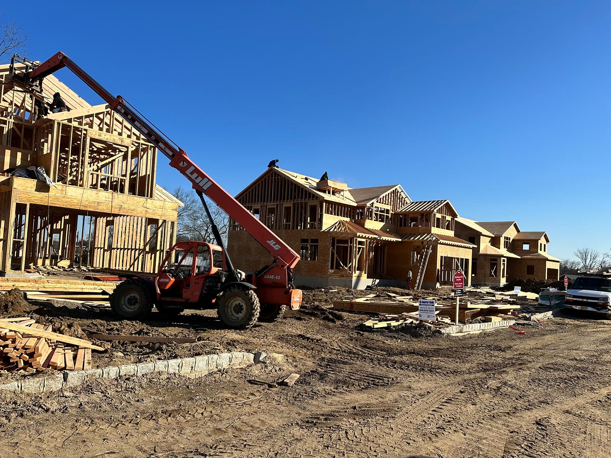 Framed two-story homes rising on a BayfordBridge parcel, telehandler lifting lumber to the roofline